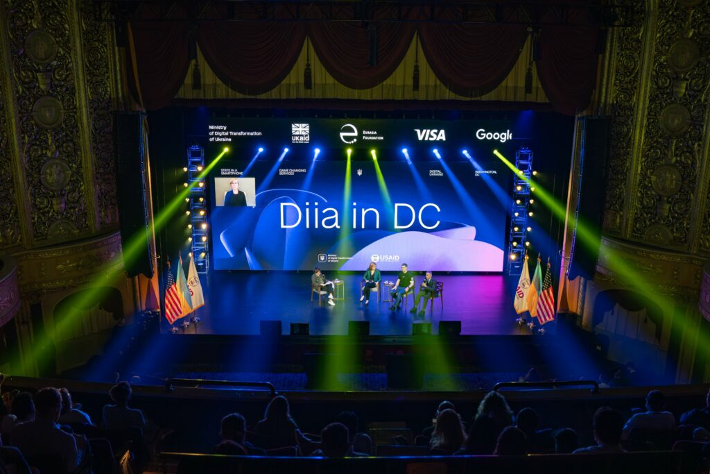 A photo shot from the second floor balcony in a theatre shows a stage with blue and yellow lights and a large backdrop reading 