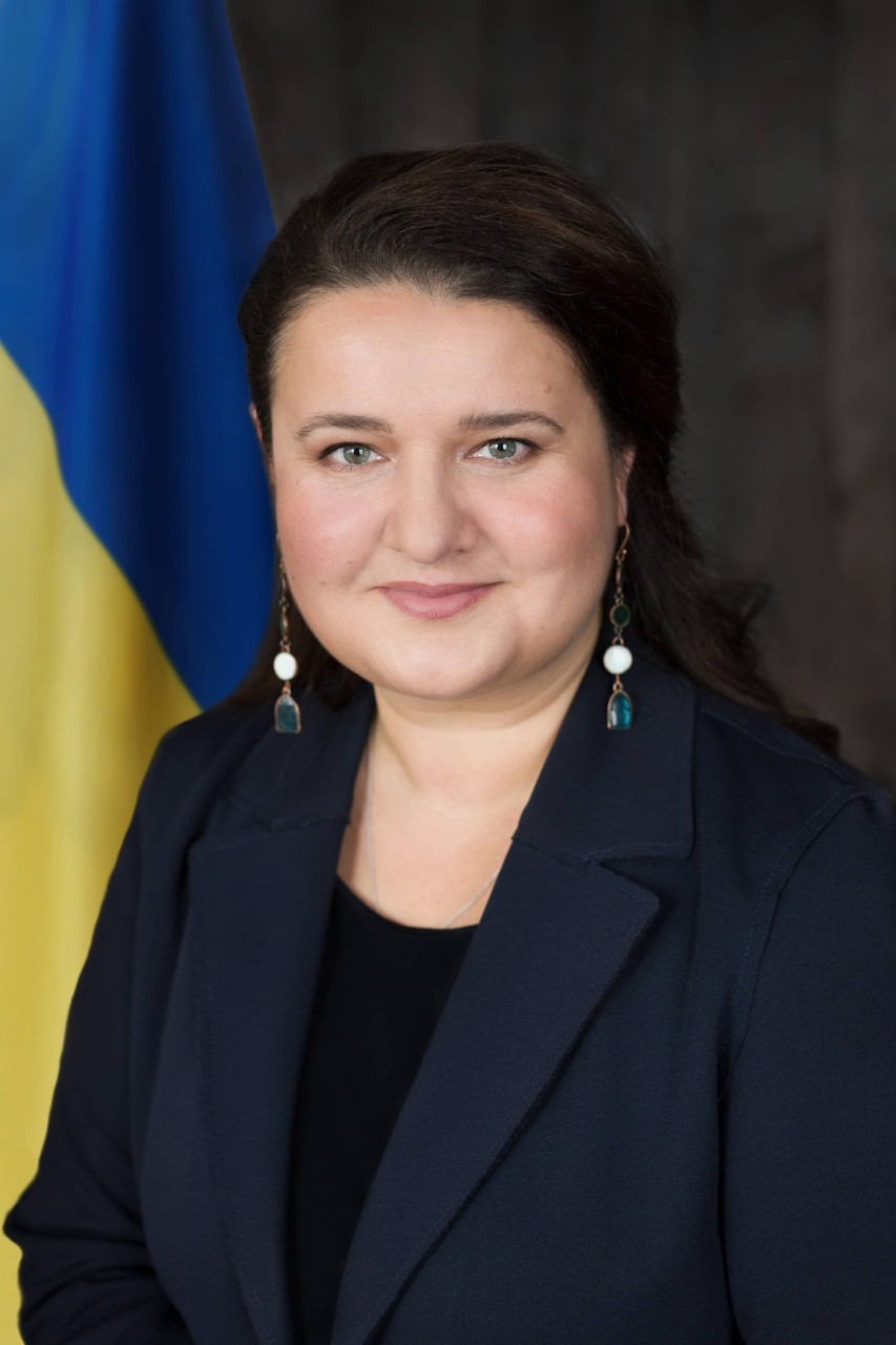 Oksana Markarova, Ambassador of Ukraine to the United States, smiles in front of the Ukraine flag.