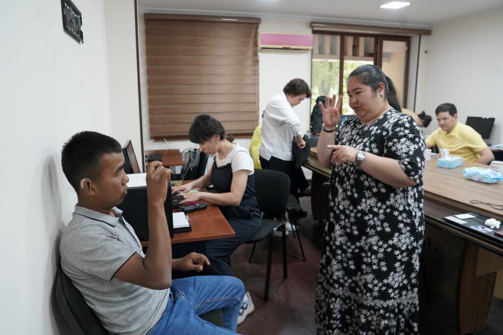 Guzal stands in an office, lifting a hand with four fingers raised. Across from her sits a man who is signing back to her with his right hand.