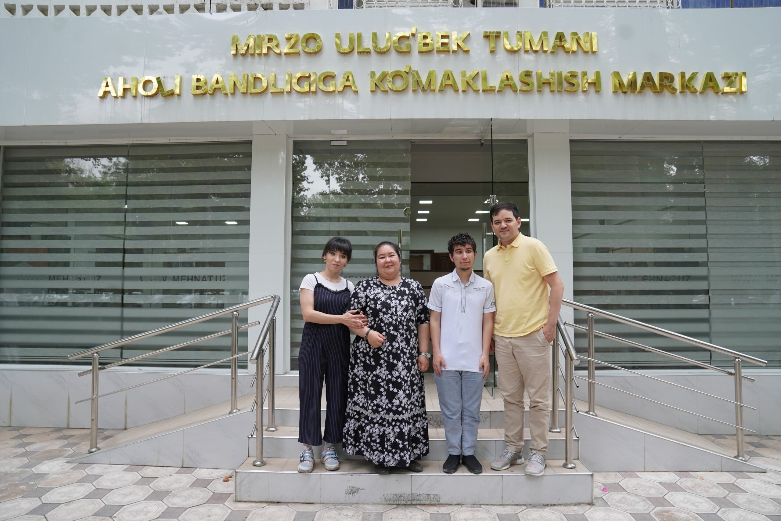 Four people--two women and two men--stand side-by-side on the steps of a gray building, smiling at the camera. The man on the right is Dilmurad.