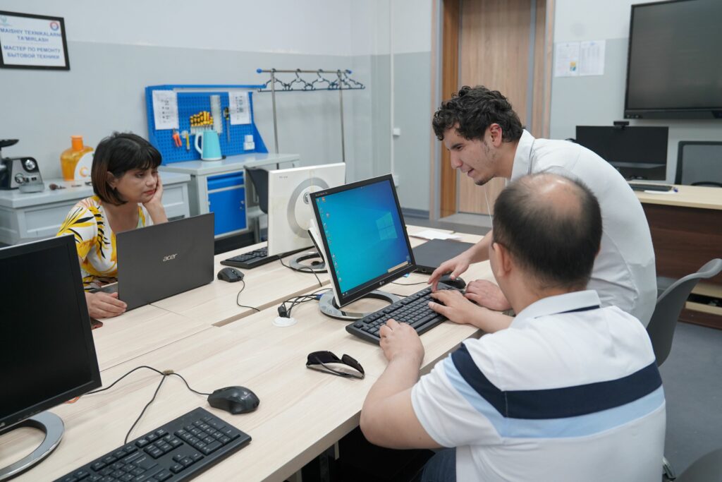 Farrukh stands at a desk with a computer in front of him. Two other people sit nearby, apparently heeding his instruction.