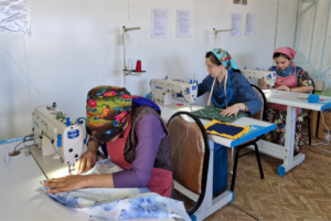 Three women in brightly colored clothing sit at individual sewing desks and use sewing machines to fashion clothing.