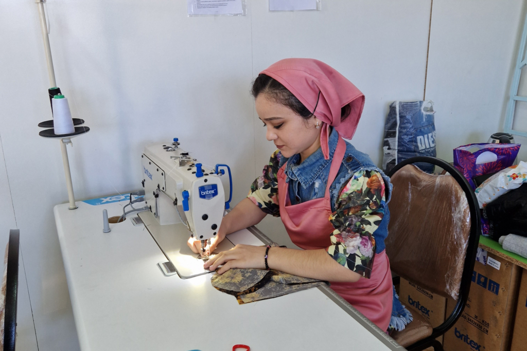 A young woman in a mulitcolor dress, pink apron, and pink headscarf operates a sewing machine at her personal desk.