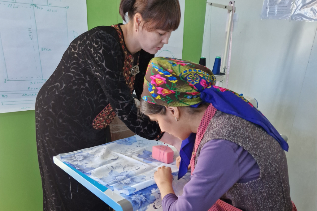 Two women lean over a desk, jointly working on a sewing project. One wears a long black dress with bright embroidery on the front and stands in front of the desk. The other woman wears a lavender dress with a green, blue, and pink floral headscarf; she sits behind the desk.