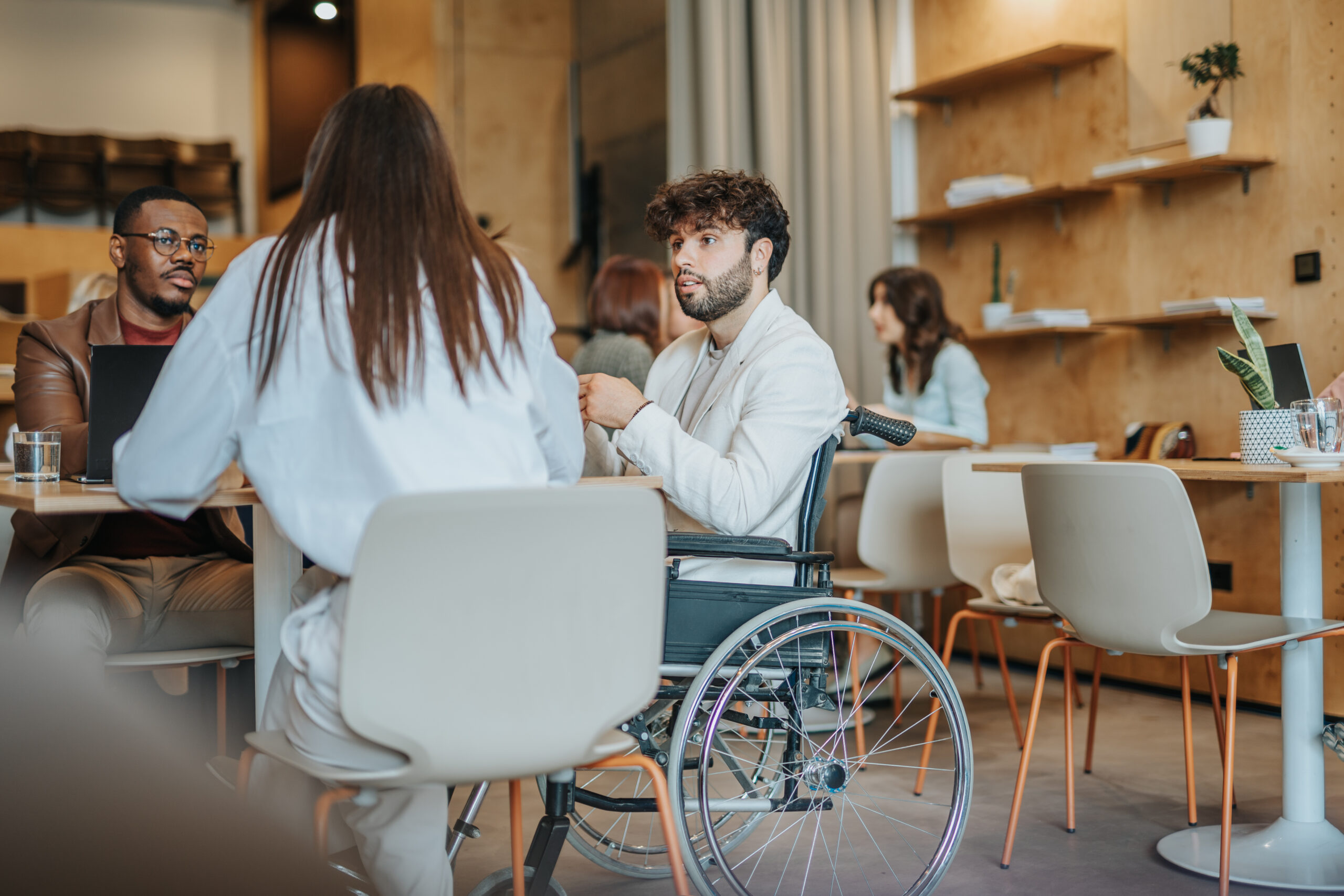 A group of three people sit around a table having a discussion. One man uses a wheelchair. They are working in a modern office or coworking space.