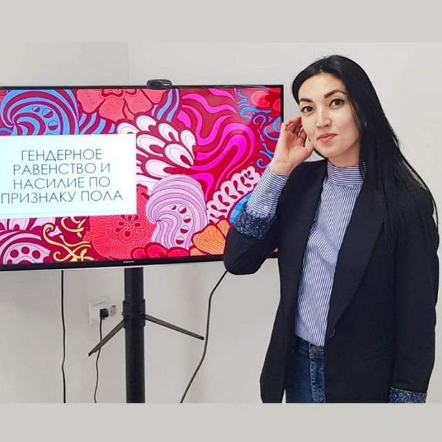 Nargiz stands in front of a TV screen displaying a presentation on gender-based violence. She looks at the camera and tucks hair behind her ear,