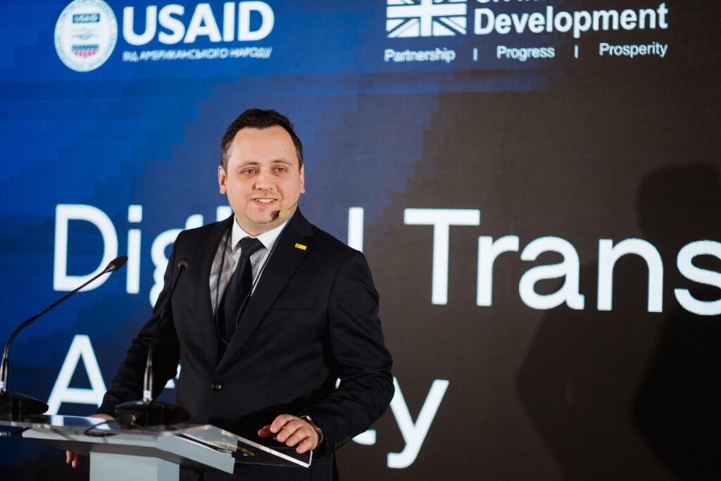 Danylo Molchanov, DTA Chief of Party, speaks at the Ukraine Digital Transformation Activity launch event. A screen behind him partially displays the program name and bears the USAID and UK Dev logos.