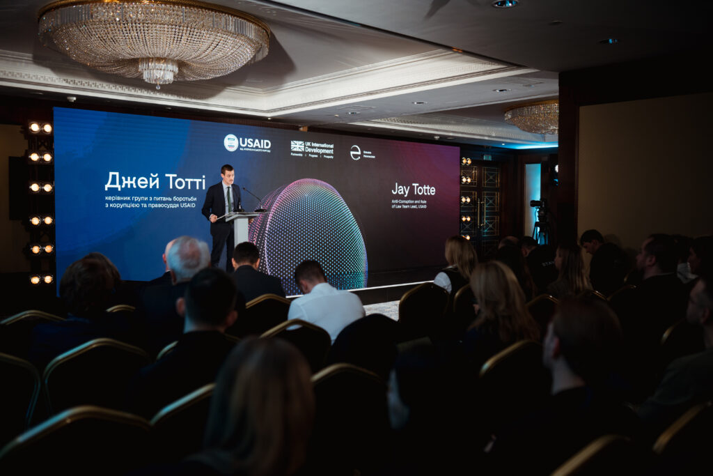 Jay Totte speaks at the Ukraine Digital Transformation Activity launch event. A large screen spanning the stage behind him shows his name in Ukrainian and English, as well as the USAID, UK Dev, and EF logos.