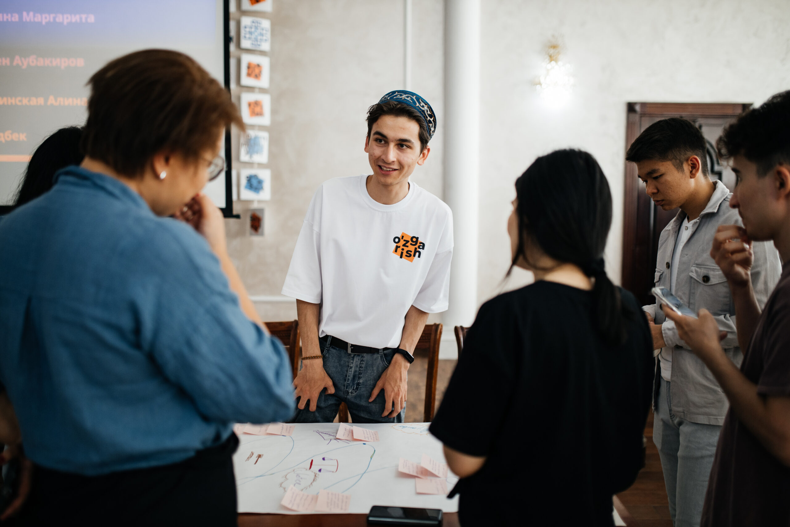 Six people gather around a table with a large sheet of paper on it, making a mind map of project ideas. The image focuses on one young man, who is wearing a CAYLA t-shirt with the Uzbek word 