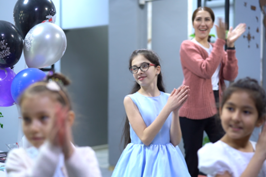 The camera focuses on a girl, approximately 8 years old, clapping along as part of a dance routine. Two younger girls stand in front of her and a teacher behind, all performing the same dance.