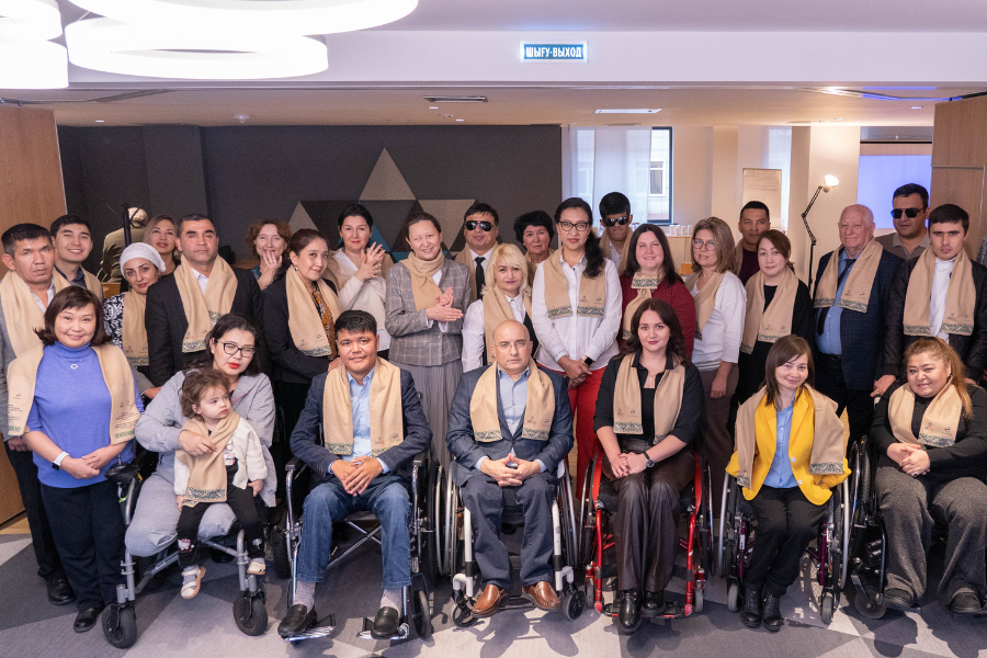A group of approximately 20 people pose and smile for a photo. Many are people with disabilities. All wear beige scarves featuring blue embroidery, the USAID logo, and the Eurasia Foundation logo.