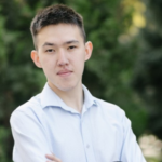 A headshot of Asylan Smagulov, a young man from Kazakhstan. He wears a professional button-up shirt and stands with his arms folded.