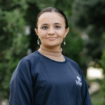 A headshot of Madina Mustafina, a young woman from Turkmenistan. She smiles at the camera and wears a navy blue Central Asia Youth Leadership Academy long-sleeve with the USAID logo over a light brown turtleneck.