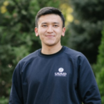 A headshot of Mahmudjon Utkurov, a young man from Uzbekistan. He smiles and sports a navy blue Central Asia Youth Leadership Academy long-sleeve with the USAID logo.