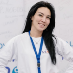 A headshot of Nargiza Rakhmanova, a young woman from Uzbekistan. She smiles at the camera with hands on her hips and wears a white Central Asia Youth Leadership Academy long-sleeve with the USAID logo.
