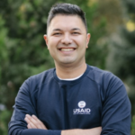 A headshot of Yusup Agajanov, a young man from Turkmenistan. He smiles at the camera with his arms crossed. He wears a navy blue Central Asia Youth Leadership Academy long-sleeve with the USAID logo.