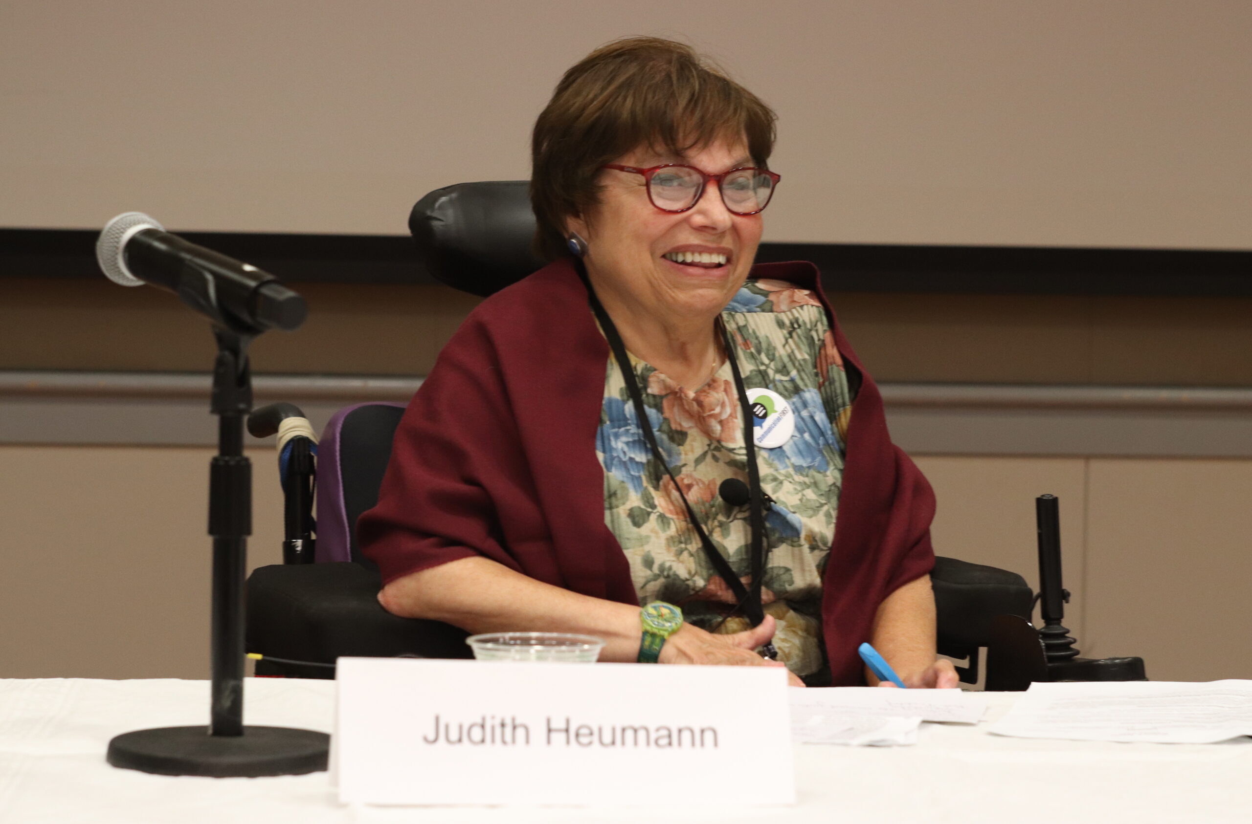 A photograph of Judith Heumann. She is sitting in her power chair behind table covered with a white table cloth. There is a microphone and a name placard in the foreground. She is wearing a colorful blouse and a maroon cardigan that matches the color of her glasses rims.