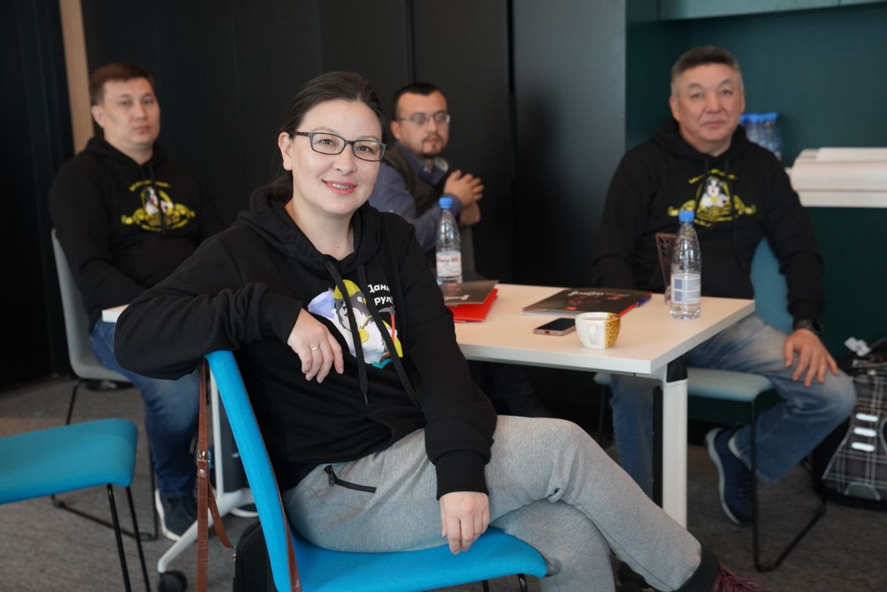 Aida Bapakhova, a co-founder of Kun Jarygy, sits at a table in a classroom with 3 other trainees. The photo focuses on Aida, sitting relaxed in her chair and smiling into the camera.