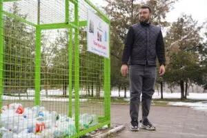 Vladislav stands to the right of a green recycling collection container. He is looking at the container sign and smiling. He is wearing a black shirt, black quilted vest, black jeans, and black shoes. There is snow on the ground behind him.