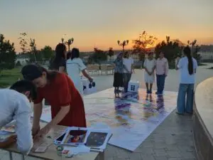 Children play table and board games in a park. In front, a boy and girl stand over a table. The table has many printouts of portrait paintings and smaller printed texts. In the back, 8 girls stand in teams on a large game board on the ground. They are playing a life-size board game with 2 large dice. One girl is holding up a sheet of paper to the other players.