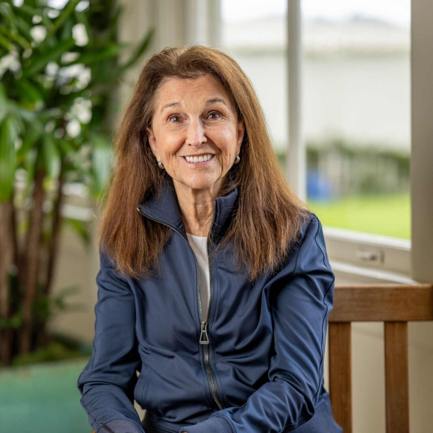 A woman with long brown hair sitting in front of a plant and a window.