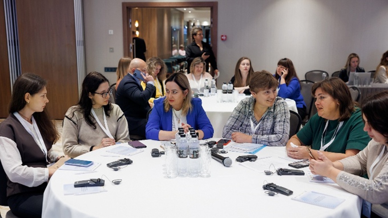 Six women sit around a circular table with a microphone in a conference room.