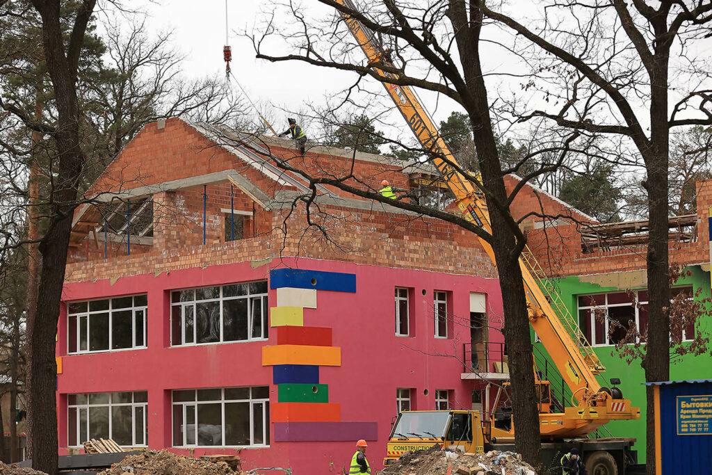 The reconstruction of a kindergarten in Ukraine. Pictured is a crane and and a worker on the rooftop of a colorful brick building that has been painted pink along with many other colors.