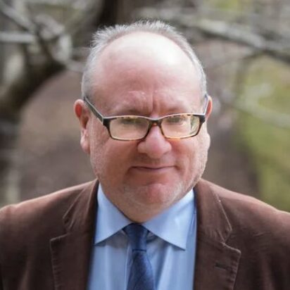 A man with glasses and a brown blazer, blue tie, and blue button-down shirt.