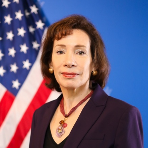 A headshot of Tracey Jacobson in a professional blazer posing in front of the U.S. flag
